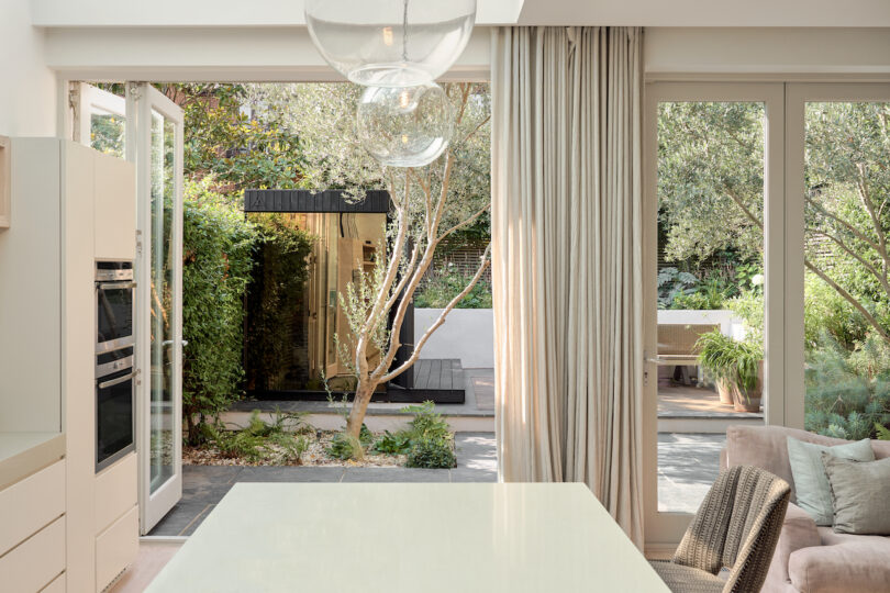 Heller kitchen area with large windows and open doors that lead to a lush garden with trees and plants. Neutral -colored curtains and furniture contribute to the airy ambience