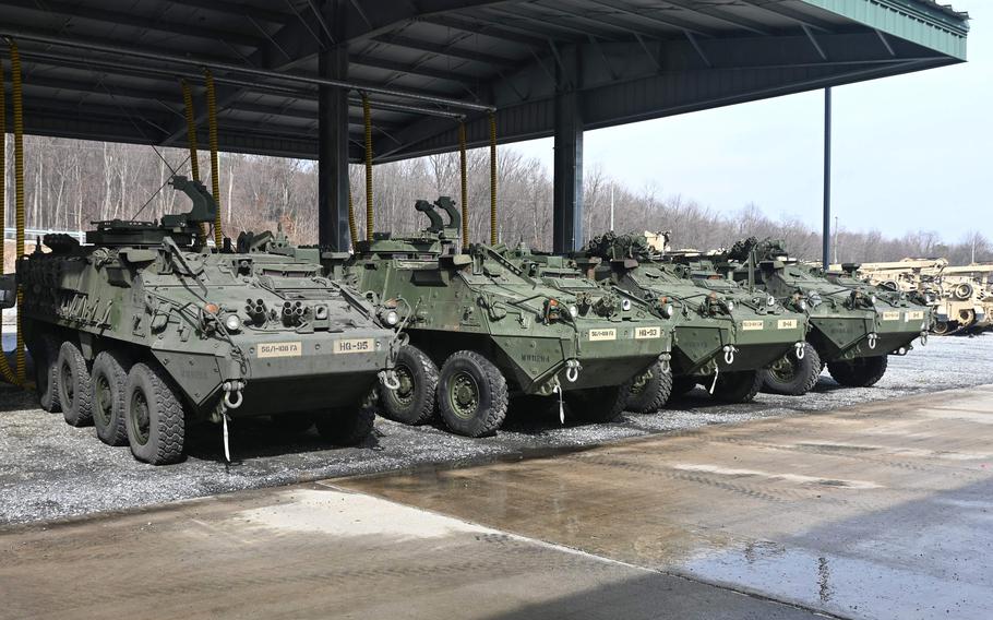 A number of four green armored vehicles rest under one roof, with other military vehicles in the background.