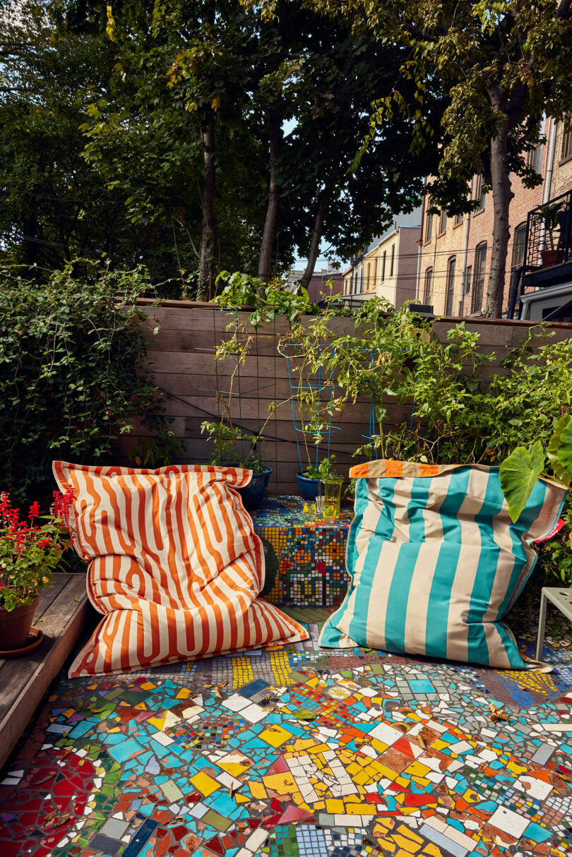 Two colorful bean bag chairs on a lively mosaic terrace, surrounded by plants and a wooden fence, with trees and buildings in the background