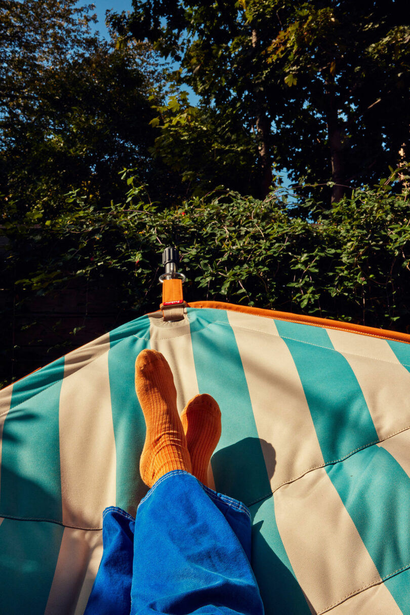 Person who relaxes in a striped hammock and wears orange socks and blue pants, surrounded by green under a clear blue sky