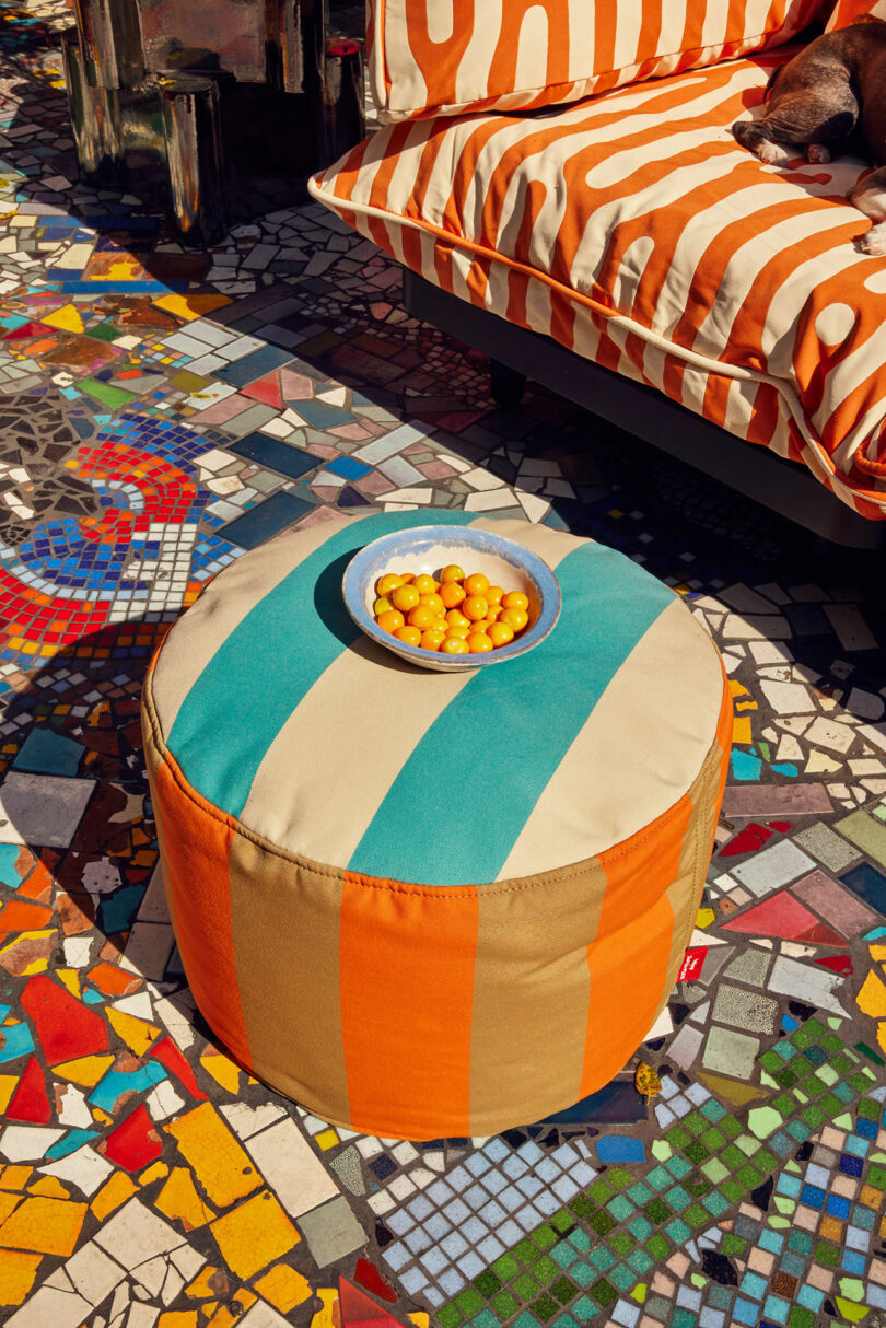 A round pouf with orange and blue -green strips keeps a bowl with small orange fruits. It is placed on a colorful mosaic floor next to a patterned couch