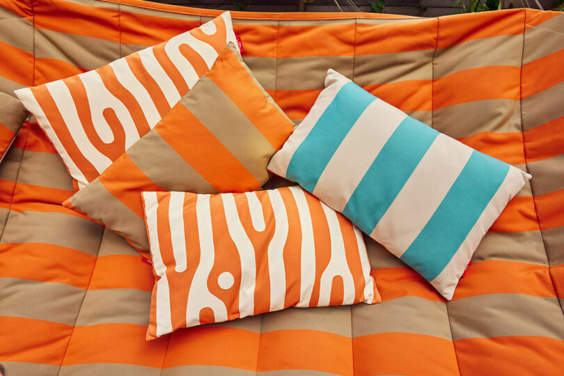 Four pillows with orange and beige patterns, including stripes and abstract designs, are placed on orange and beige striped padded surface