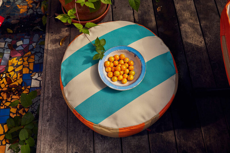 A round pouf with orange and blue -green strips keeps a bowl with small orange fruits.