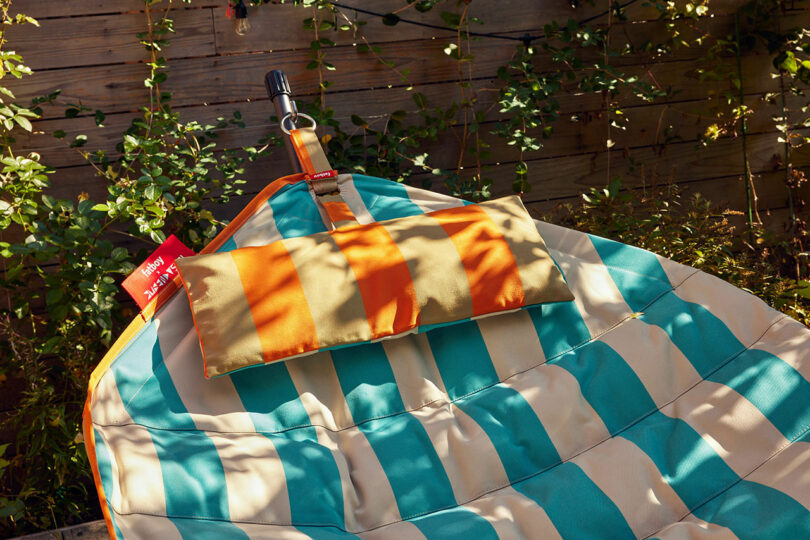 Striped outdoor hammock with blue and beige fabric with an adjustable orange headrest is furnished between wooden posts with surrounding green