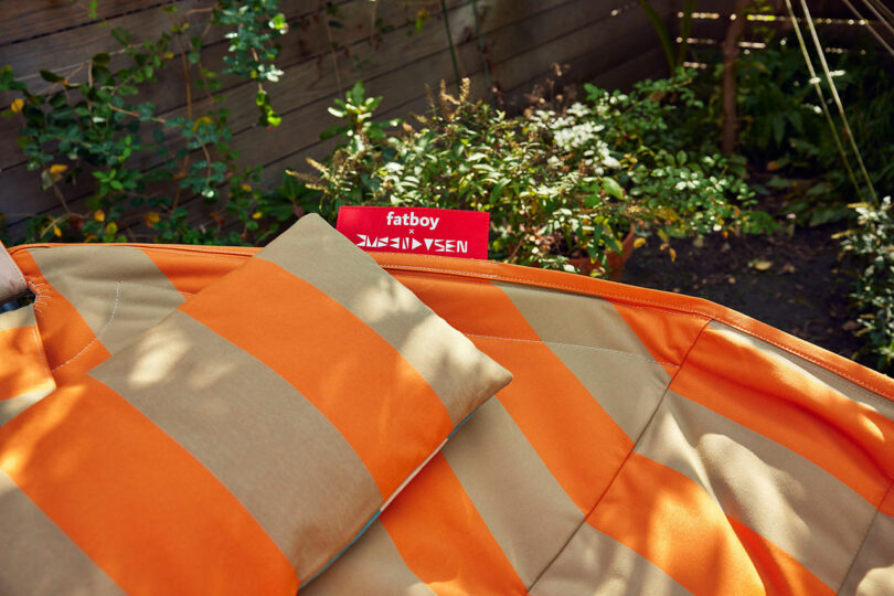 A light orange and beige striped outdoor pillow with a red "Fatboy" Day rests on a garden hammock with green in the background