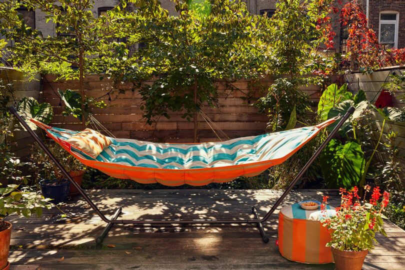A colorful striped hammock is hung on a metal frame in a lush garden garden, surrounded by plants and wooden fences, with a suitable round pillow on the deck