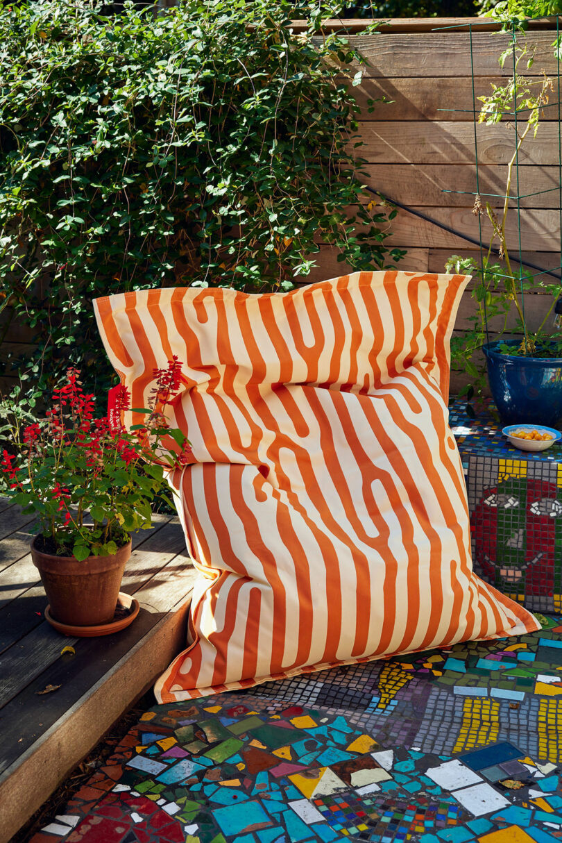 Large orange and white striped pillows on a mosaic tile terrace next to a potted plant and a wooden fence with green