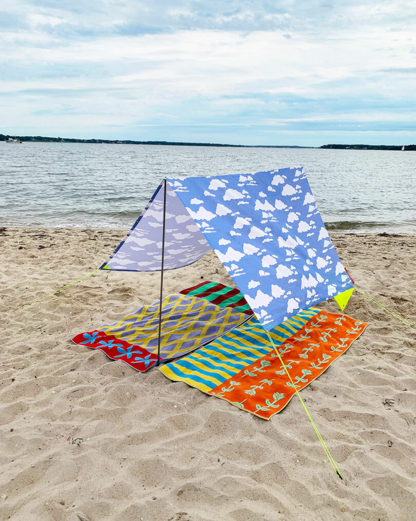 A beach setup with a blue -white canopy with cloud patterns that cover colorful mats on the sand near a quiet water under a cloudy sky