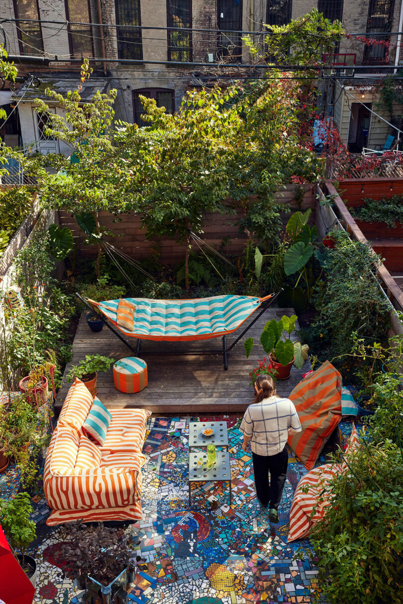 One person stands on a colorful mosaic terrace with striped outer furniture and a hammock, surrounded by lush green and high plants