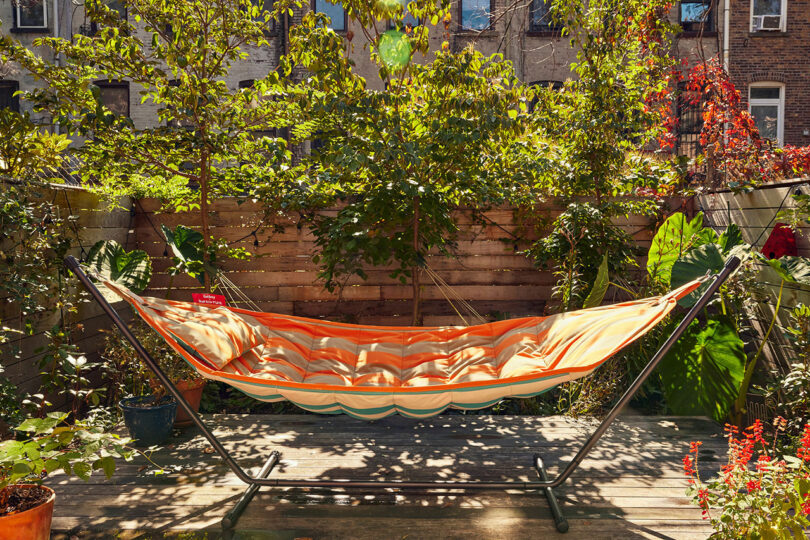 A striped hammock is hung on a metal frame in a sunlit garden, surrounded by leafy plants and a wooden fence