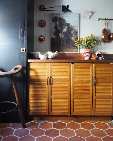 Wooden cabinet with upscale doors, copper pots, a flower vase and a painting on the wall in a kitchen.