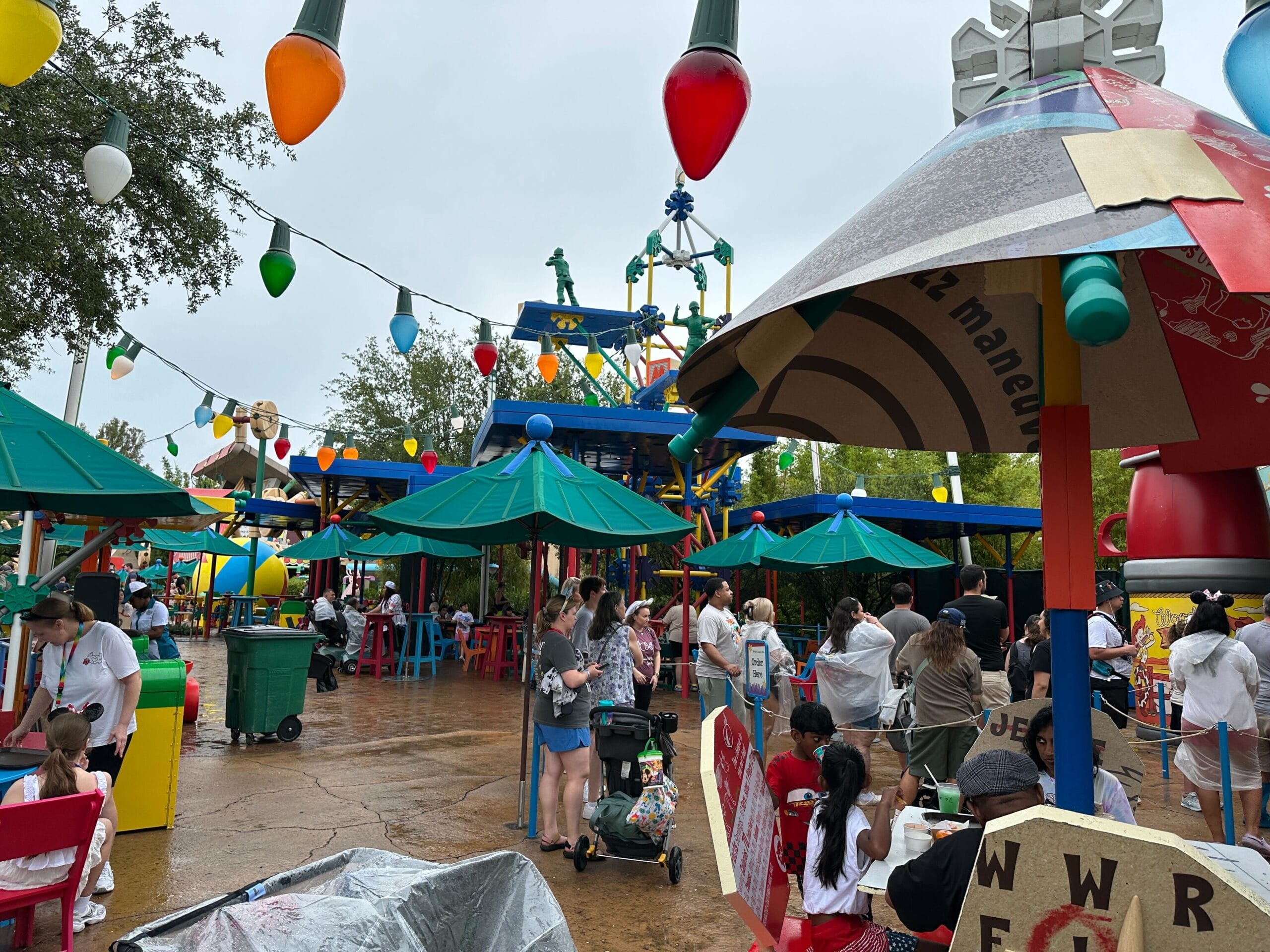 People go and sit under umbrellas in a colorful outdoor area with toy motifs, which is decorated with huge string lights.