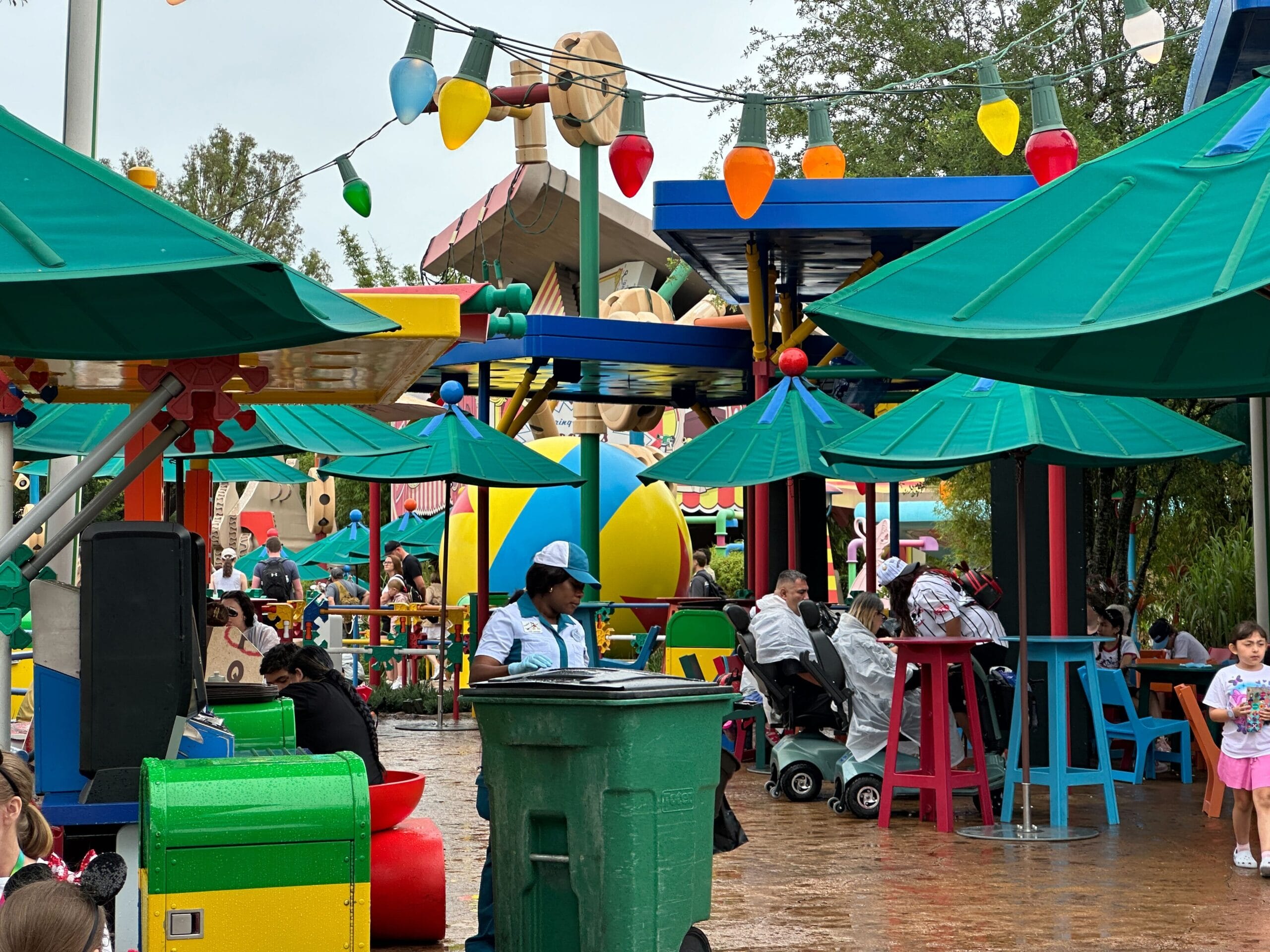 Colorful outdoor seating area with green umbrellas, people and large decorations with toy motifs in a theme park.