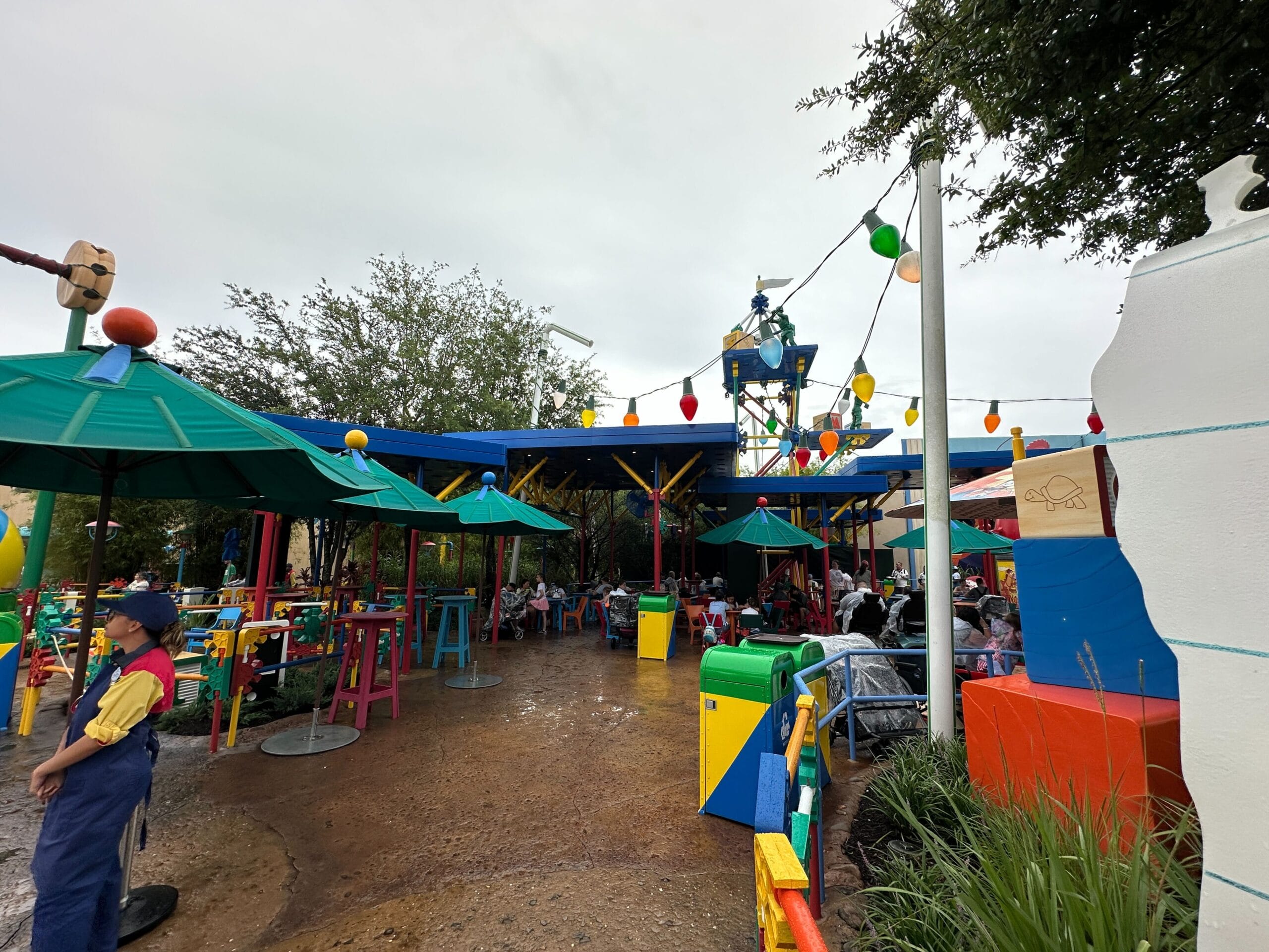 Outdoor dining area with colorful tables, umbrellas, string lights and people sitting under a cloudy sky.