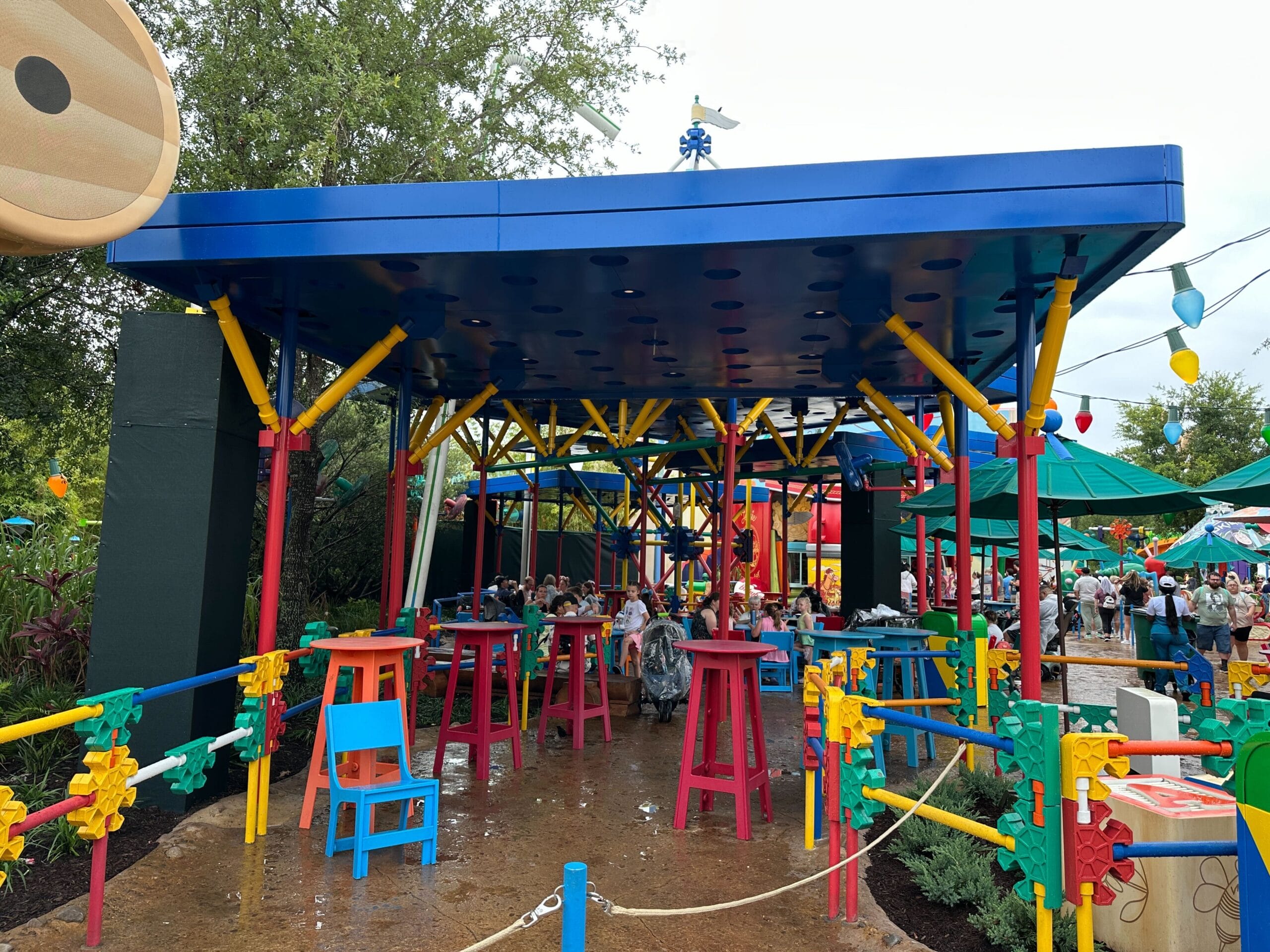 Outdoor seating area with colorful tables, chairs and structures inspired with toys, surrounded by green and playful decor.