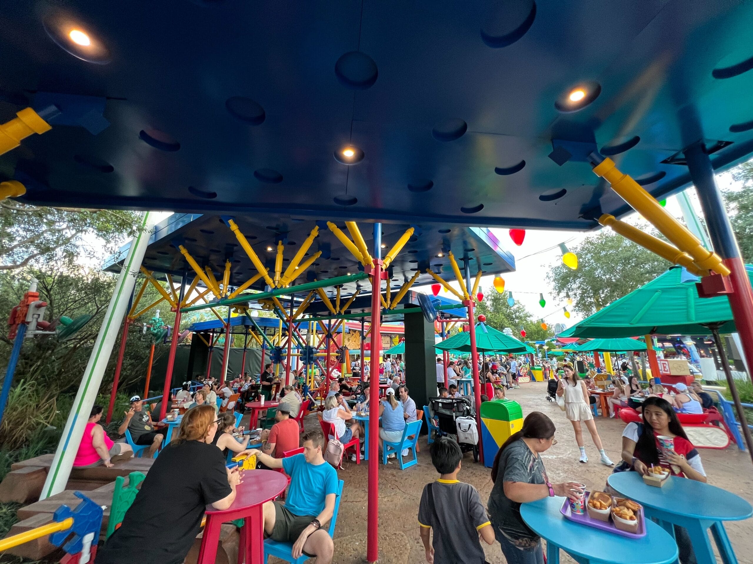 People outdoors of colorful tables under a blue canopy, which is adorned with playful, oversized, excessive toy structures.