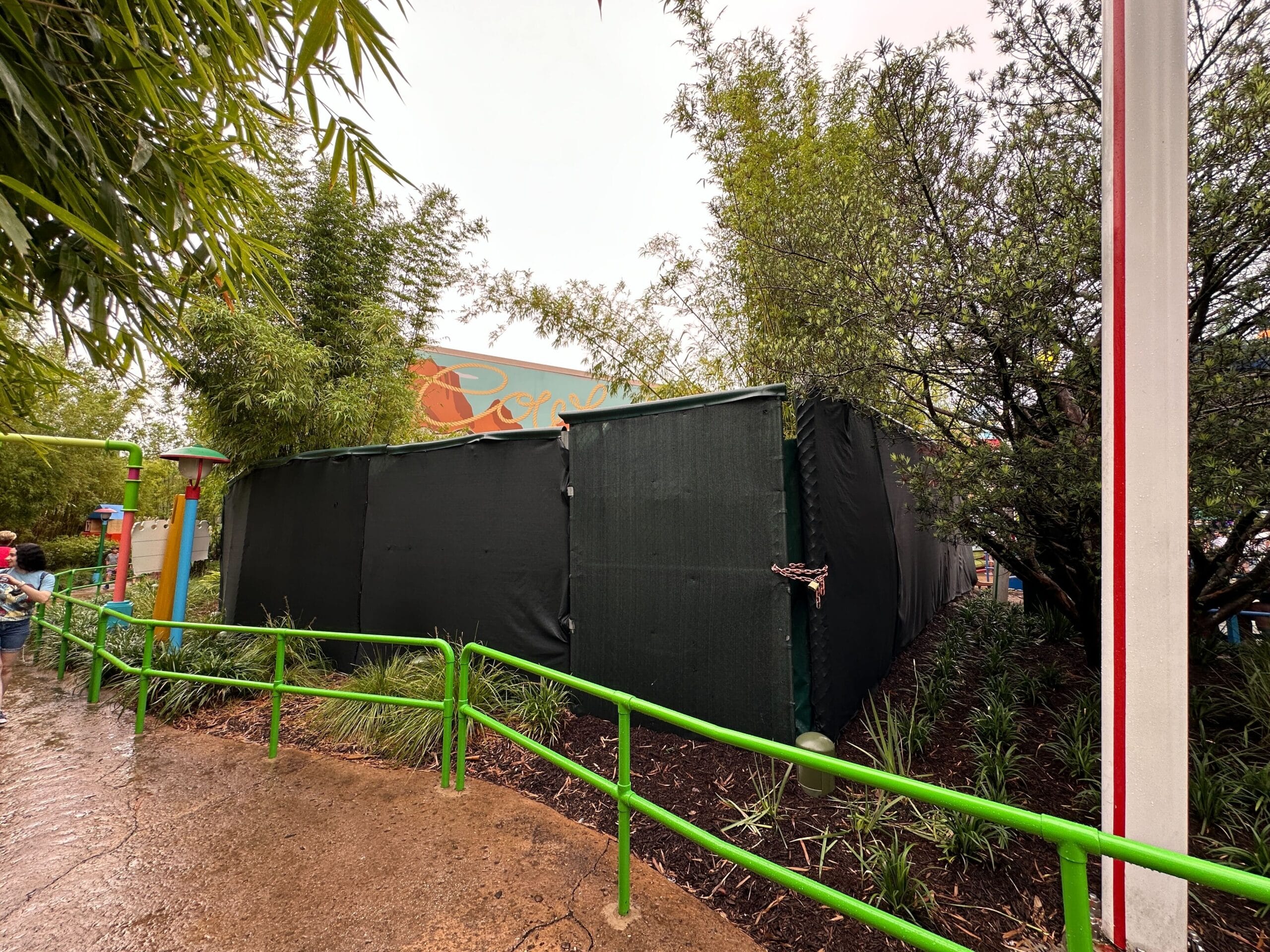 Green construction walls block an area near plants and a sign with a rain path and a green railing.