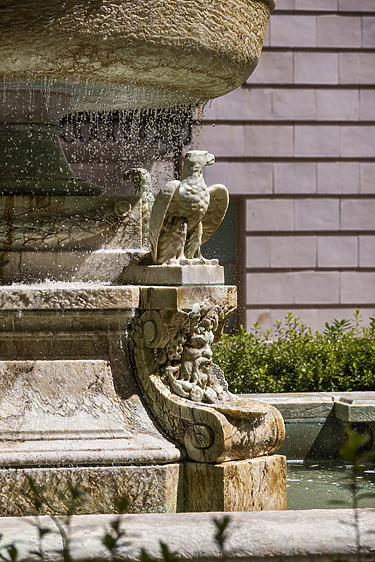 Stone sculpture of an eagle on a decorative fountain edge with water, surrounded by green bushes and a wall.