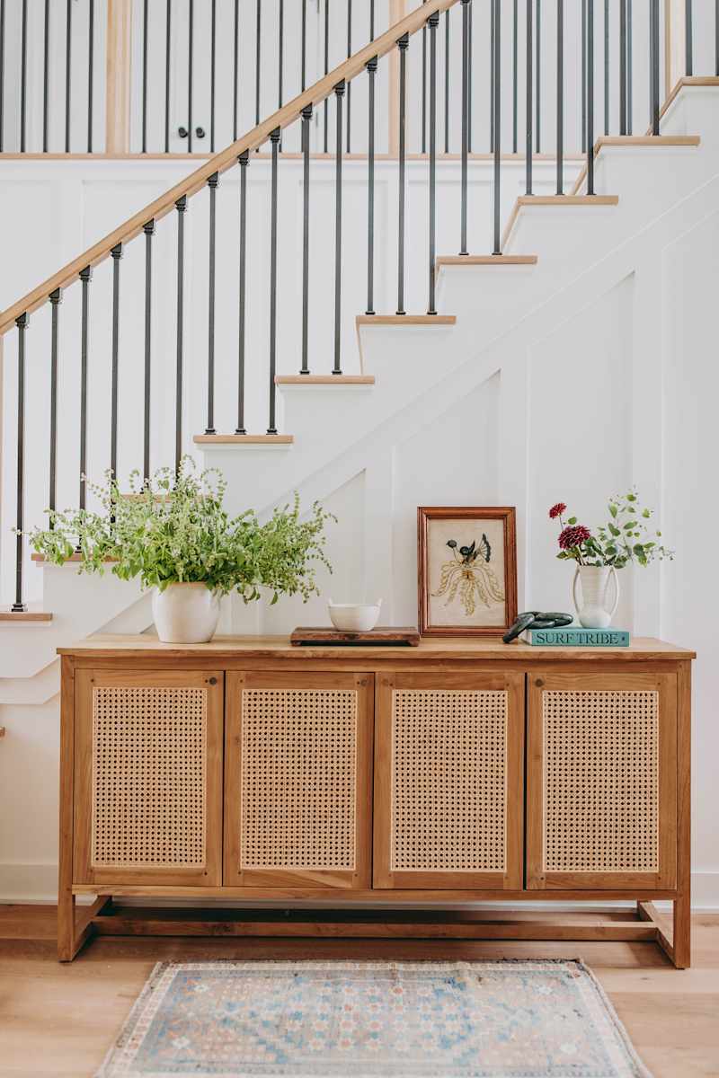 Wood sideboard with sugar cane doors, potted plants, framed artwork and decorative books under a staircase.