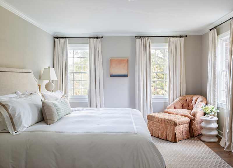 Light bedroom with a large bed with white bed linen, a patterned armchair and light curtains that frame the windows.