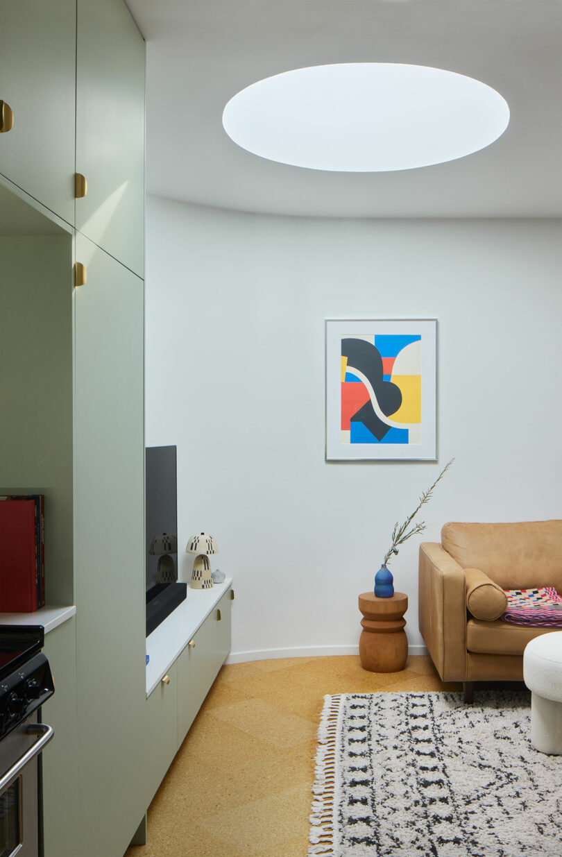 A modern living room with a round skylight, abstract art on the wall, a beige sofa, a patterned carpet and a television on a white cabinet.