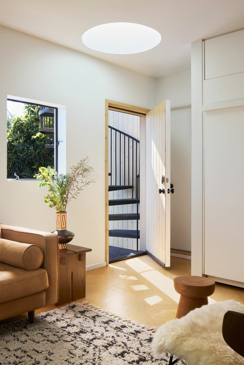 A modern living room with brown sofa, patterned carpet and wood accents; The open door shows a spiral staircase and a green outdoor.