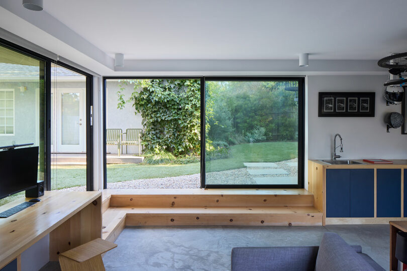 Modern living area with large glass sliding doors with a view of a green garden with wooden benches, a desk, a small kitchenette and minimalist decor.