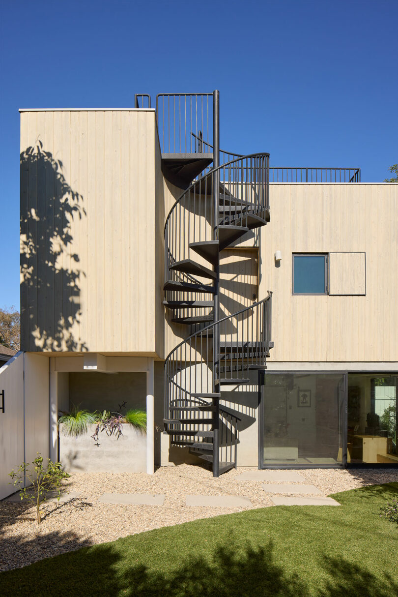 A modern two-story building with a bright wooden oxygen has a black metal staircase that leads to a roofing cover that plays in a scenic garden with grass and gravel.