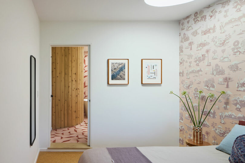 A modern bedroom with patterned wallpaper, a bed, a flower vase, two framed pictures on the wall and an open door that shows a wood -covered room with a geometric tile floor.