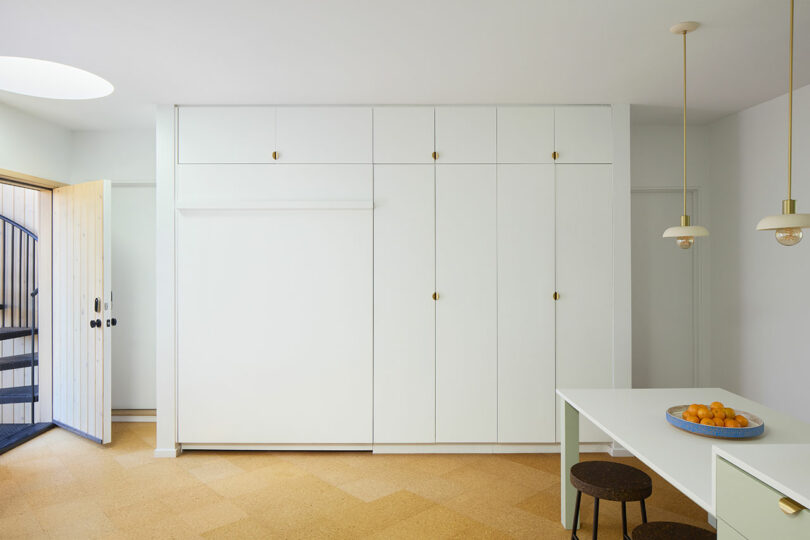 Minimalist cuisine with white built -in cupboards, a table with chairs, trailer lights and a bowl of oranges on the table. A door with a safety goal is open on the left.