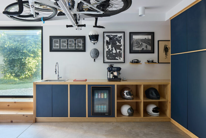 Modern room with blue and wooden cabinets, a small sink, coffee machine, mini cooler, bicycle helmets and bicycle that are mounted on the ceiling; Framed art hangs on the wall.