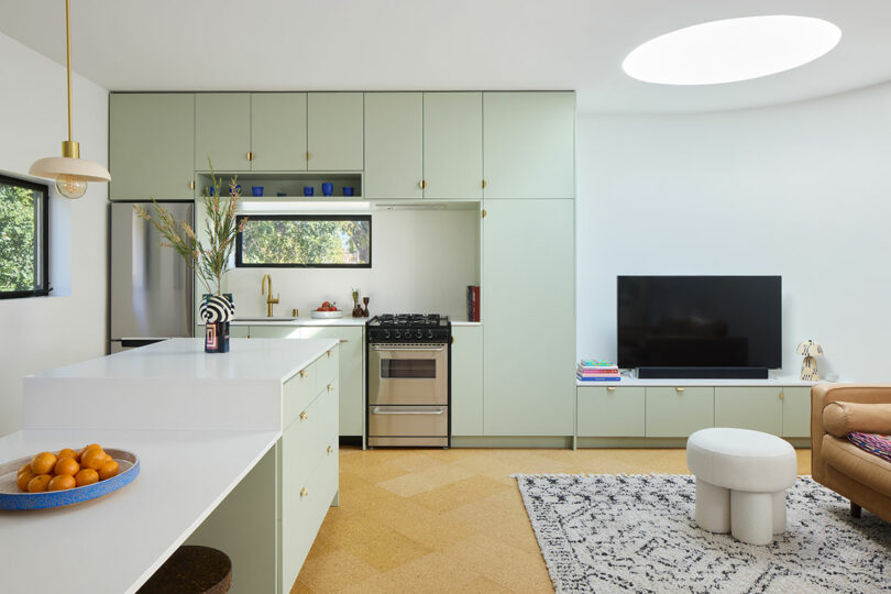 Modern kitchen and living areas with light green cupboards, stainless steel devices, a television mounted on the wall, a round Ottoman and a bowl orang on a white island.