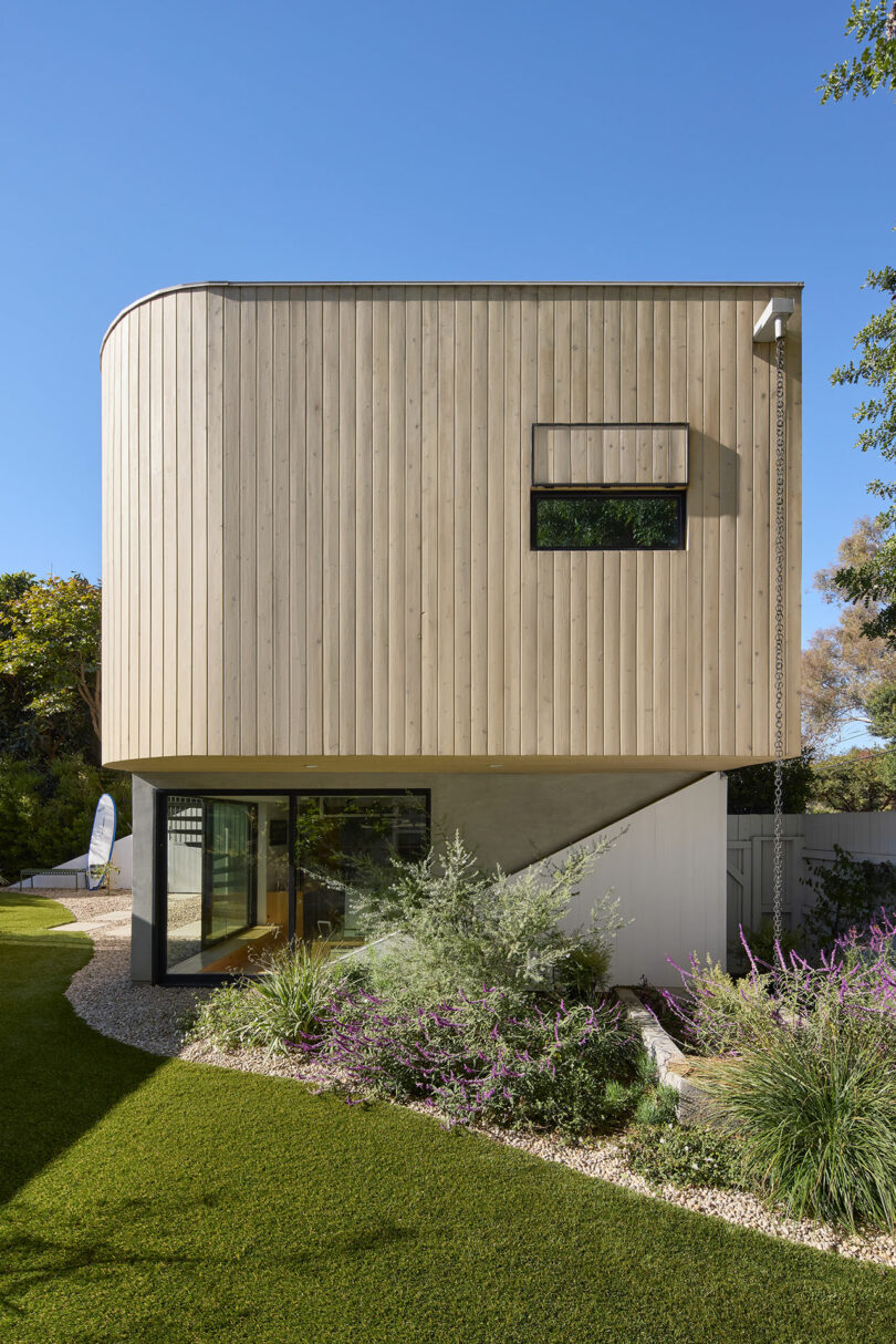 Modern two -story house with a curved wooden upper facade, a large glass of the lower level and a created garden with green grass and shrubs.