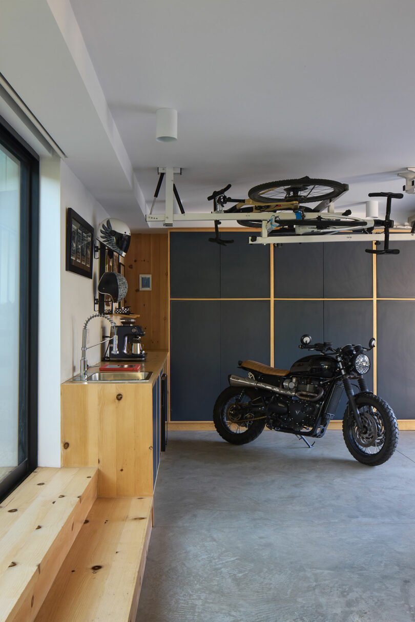 A modern garage with a motorcycle that is parked on a concrete floor, a bike over your head, wooden cabinets and a worktop with a sink and coffee machine.