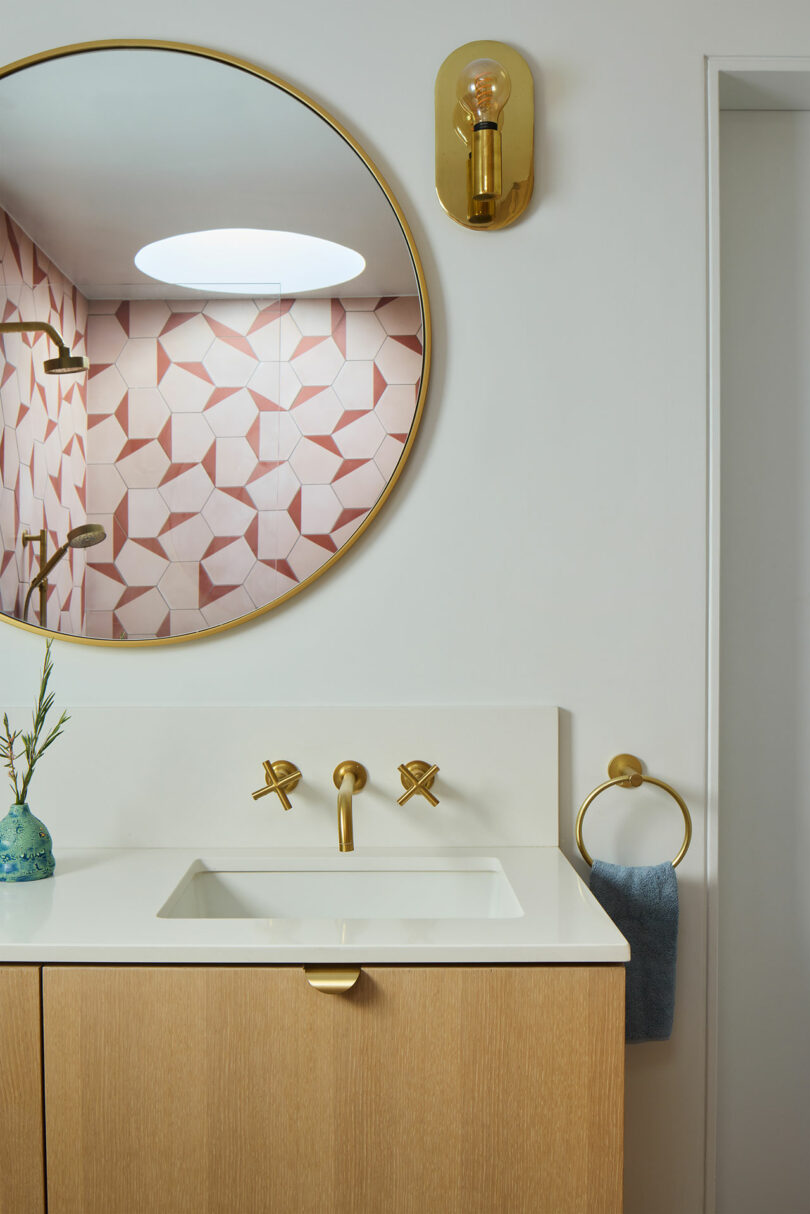 Modern bathroom with a round mirror, brass fittings, light wood washbasin, white worktop and geometric pink tiles that are reflected from the shower.