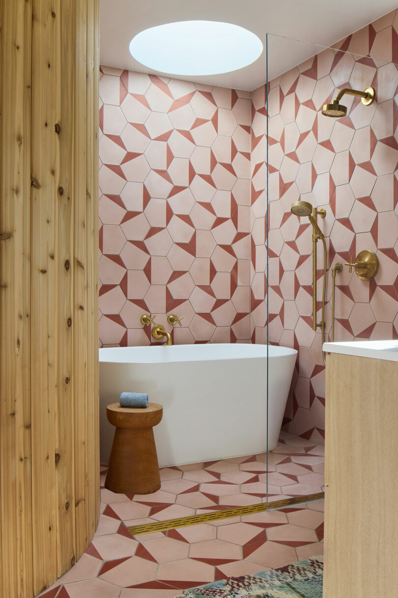 Modern bathroom with geometric pink tiles, a white free -standing tub, gold lights, a wooden stool and natural wood accents under a round skylight.