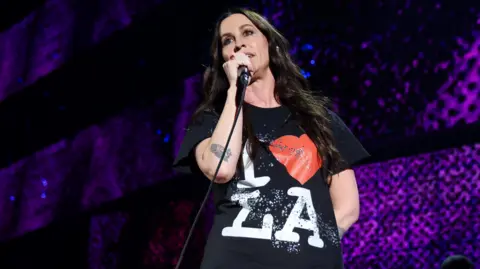 Getty pictures A woman with brown hair smiled in the camera black top. She holds a microphone with the stage design behind her, indicating that she is in a live performance.