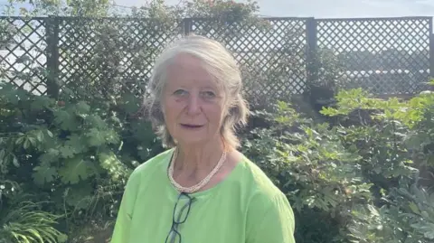 A woman who wears a green dress is in a garden with bushes and a grille behind her. She has a golden necklace and her glasses hang on the top of her dress.