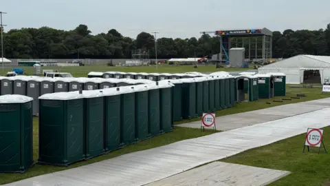 A series of portaloos can be seen in front of a stage that is built on the blackweir name connected to a state. There is also a white tent on the right in the photo. They are plastic paths in the grass.