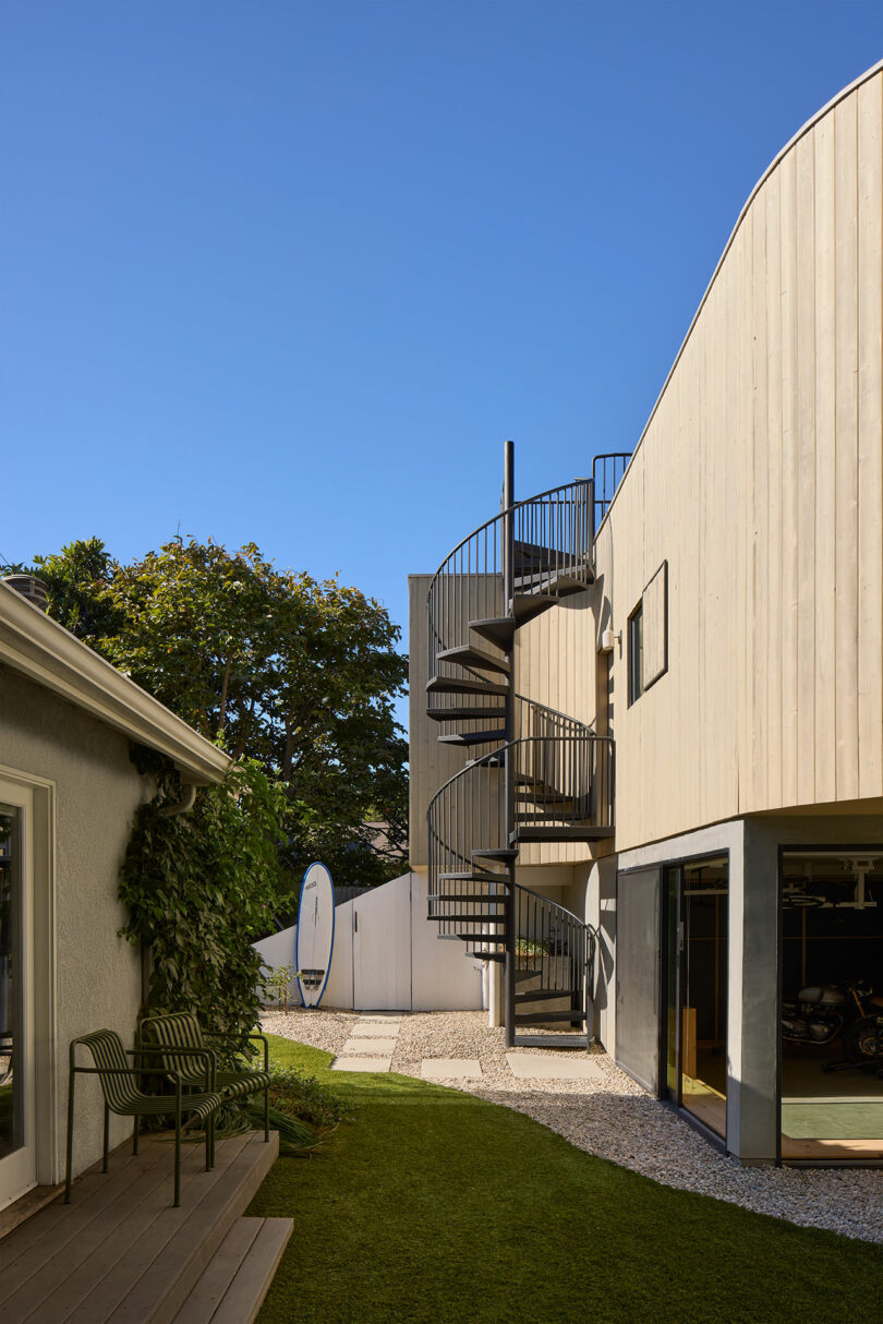 A modern building with a light wood cladding area has an outer metal staircase, next to a lawn and another structure under a clear blue sky.