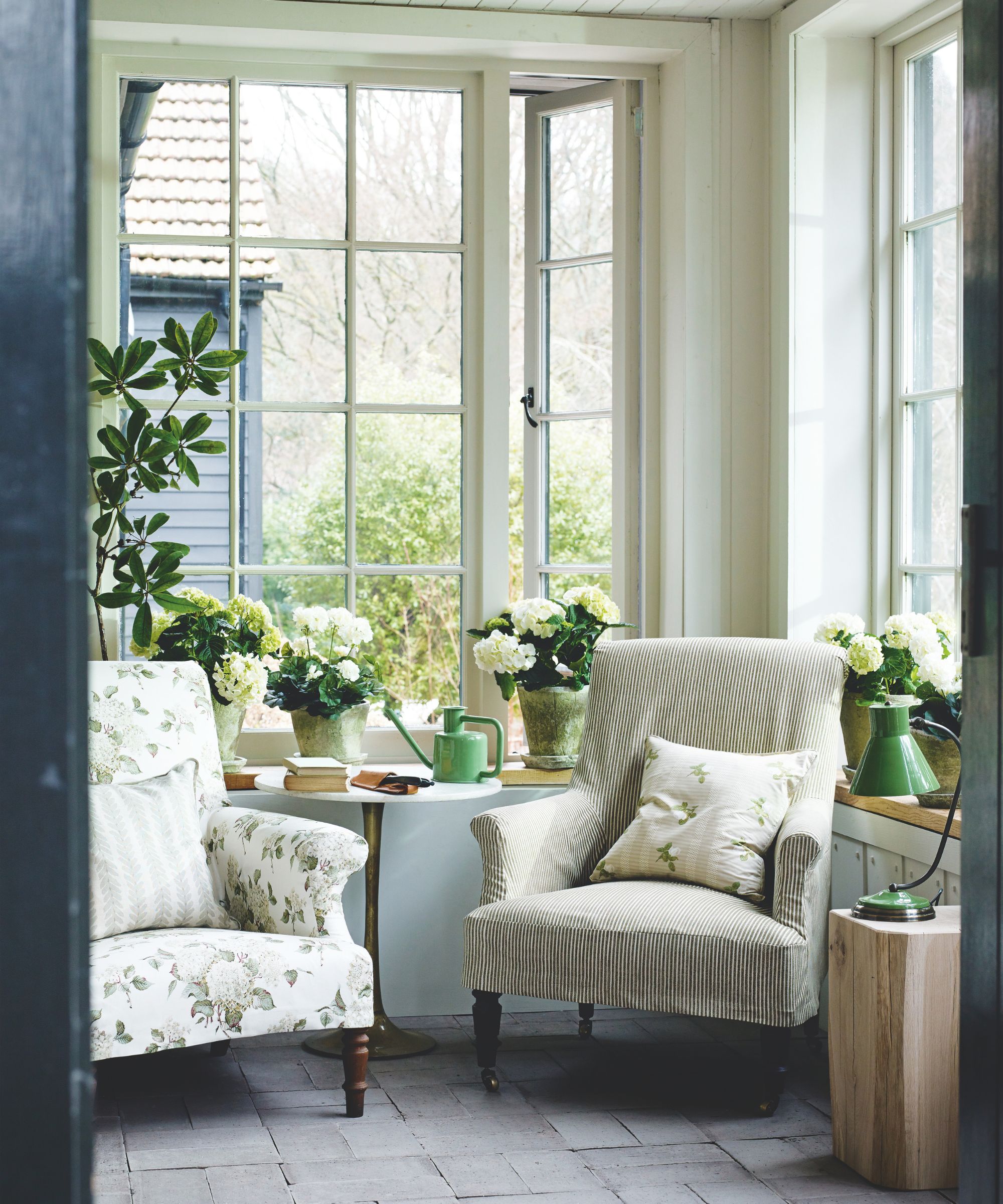 A bright and sunny winter garden with two floral and striped white and green armchairs. Behind the chairs there are large open windows and houseplants in green pots. To the right of them is a wooden side table with a green lamp. The flooring is gray and tiled.