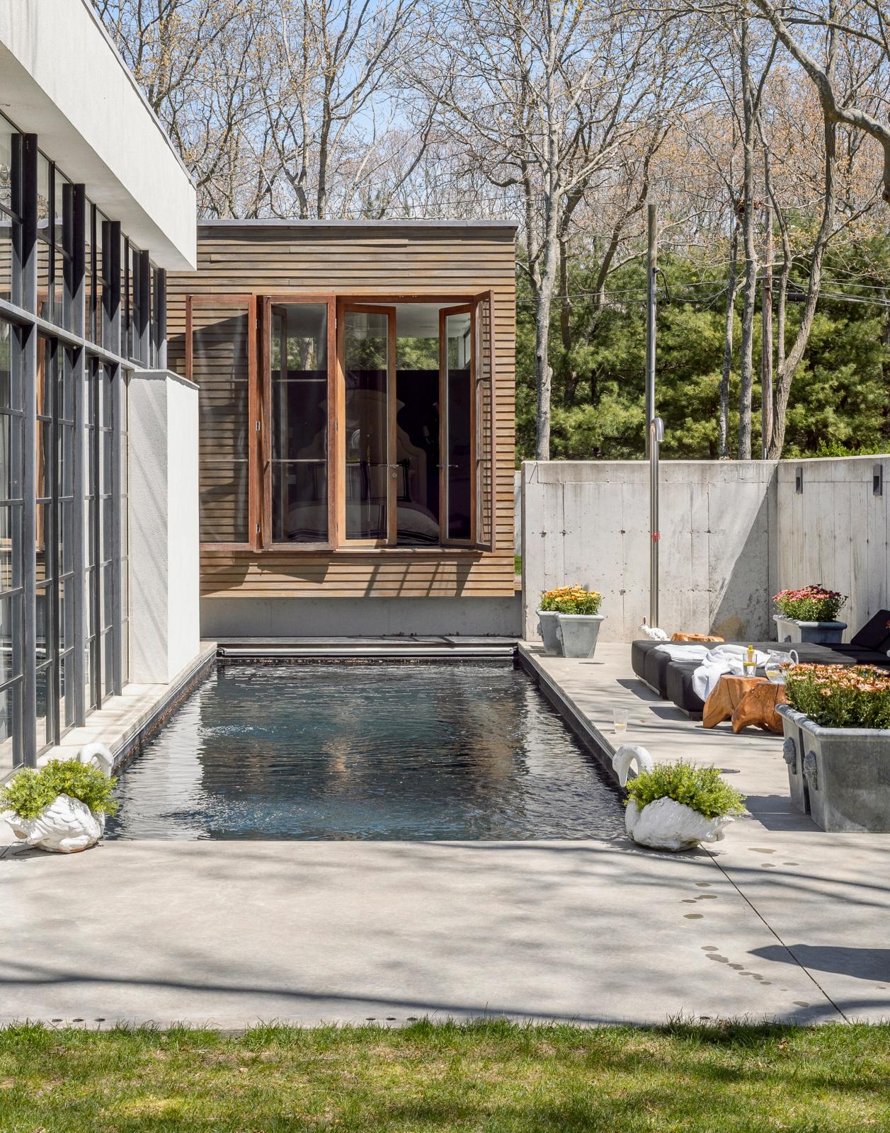 Image of a pool area outdoors with the corner of a house around him. The walls of the house are white and wood with many windows that reflect the light.