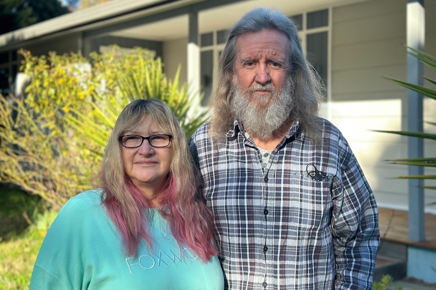 A woman with long blond hair with pink ends and a man with long gray blonde hair and a beard stand in front of a house.