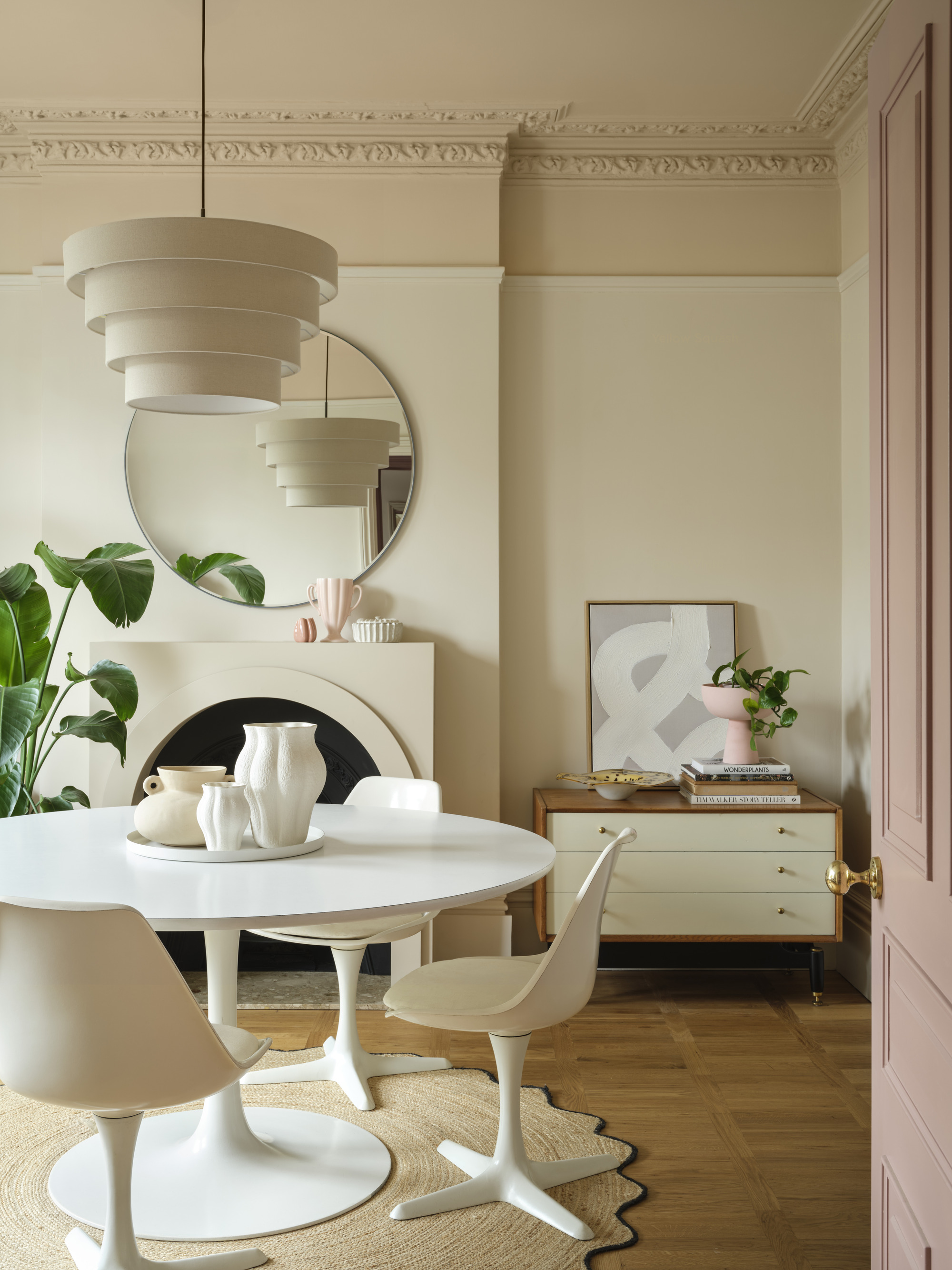 Image of a dining room that was painted in different cream and beige with a color-capper color technique. There is a modern round, white dining table and suitable modern chairs. Under the table there is a baked jute carpet and a fireplace on the back wall.