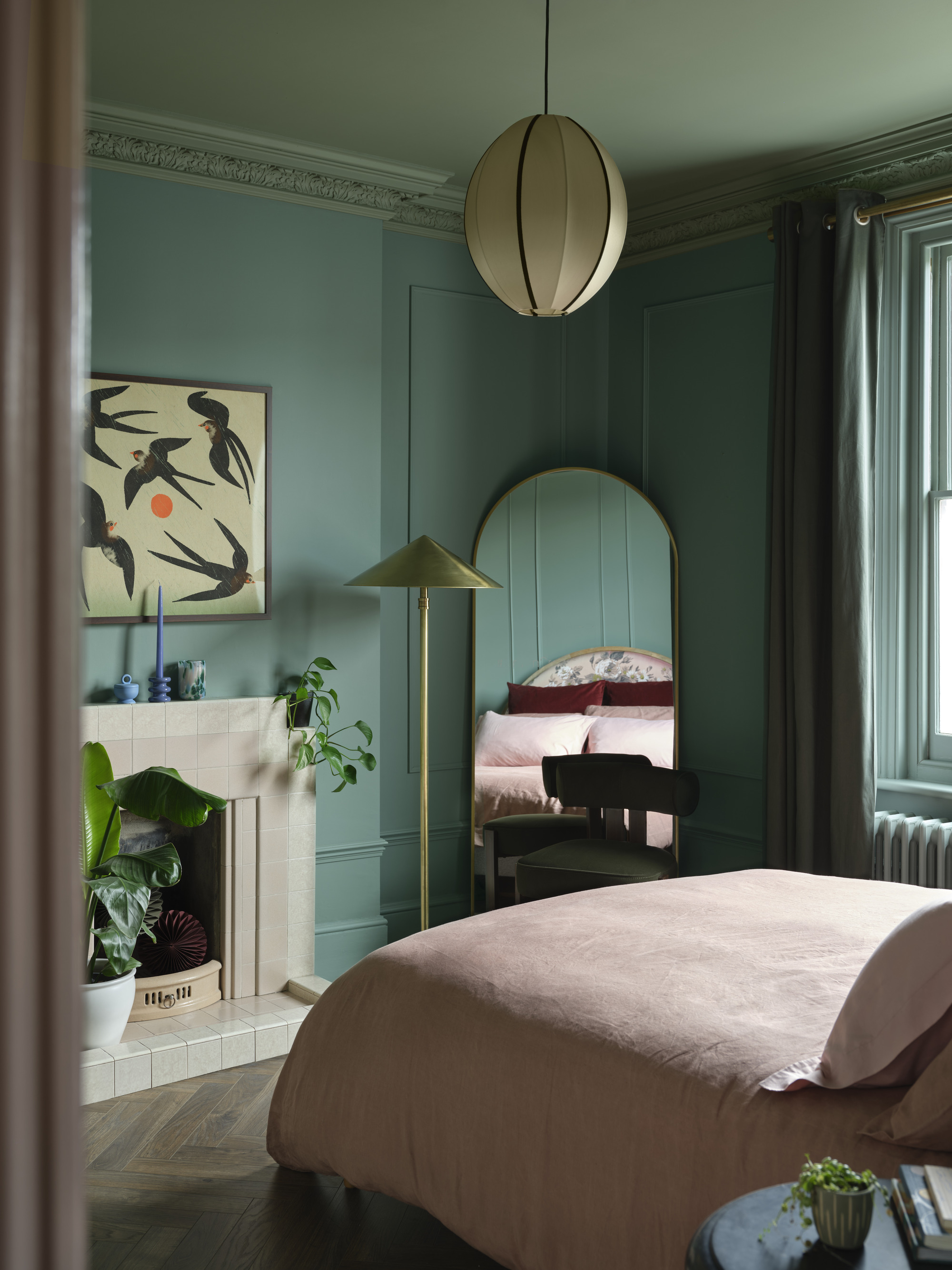 Image of a bedroom in which the walls are painted in a color cap technique using different shades of dark blue -green color. There is a bed with dusty pink bed linen and a matt -long mirror in the corner.