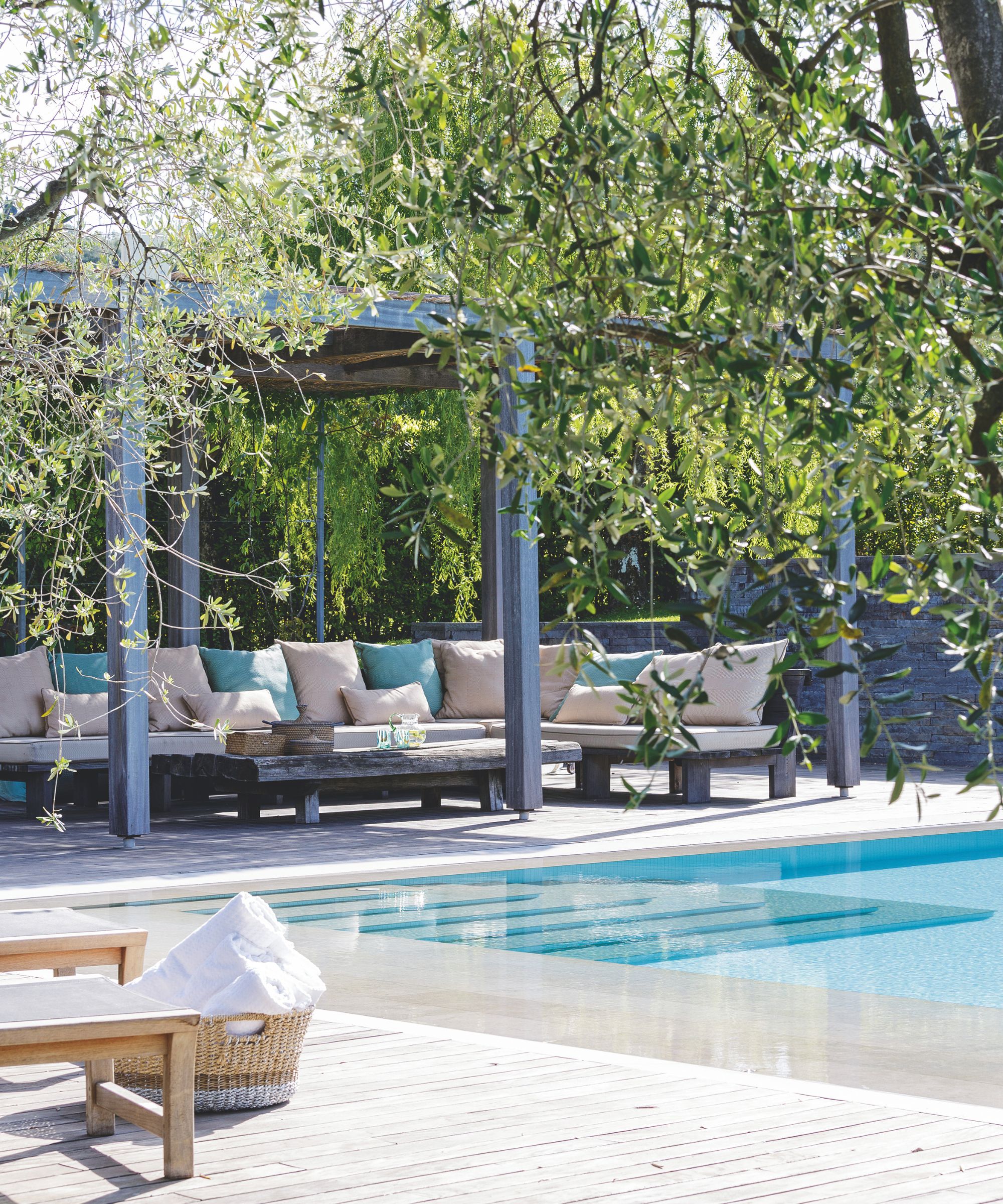 A leaves and sunny pool area with a slightly washed deck, sun lounger with a basket basket with white towels and a large seating area with pink and blue pillows under a covered canopy under the trees.