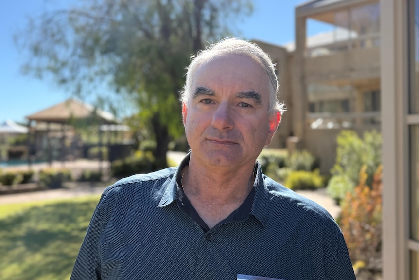 An older man who is bald with dark, bushy eyebrows has a neutral expression in the direction of the camera.