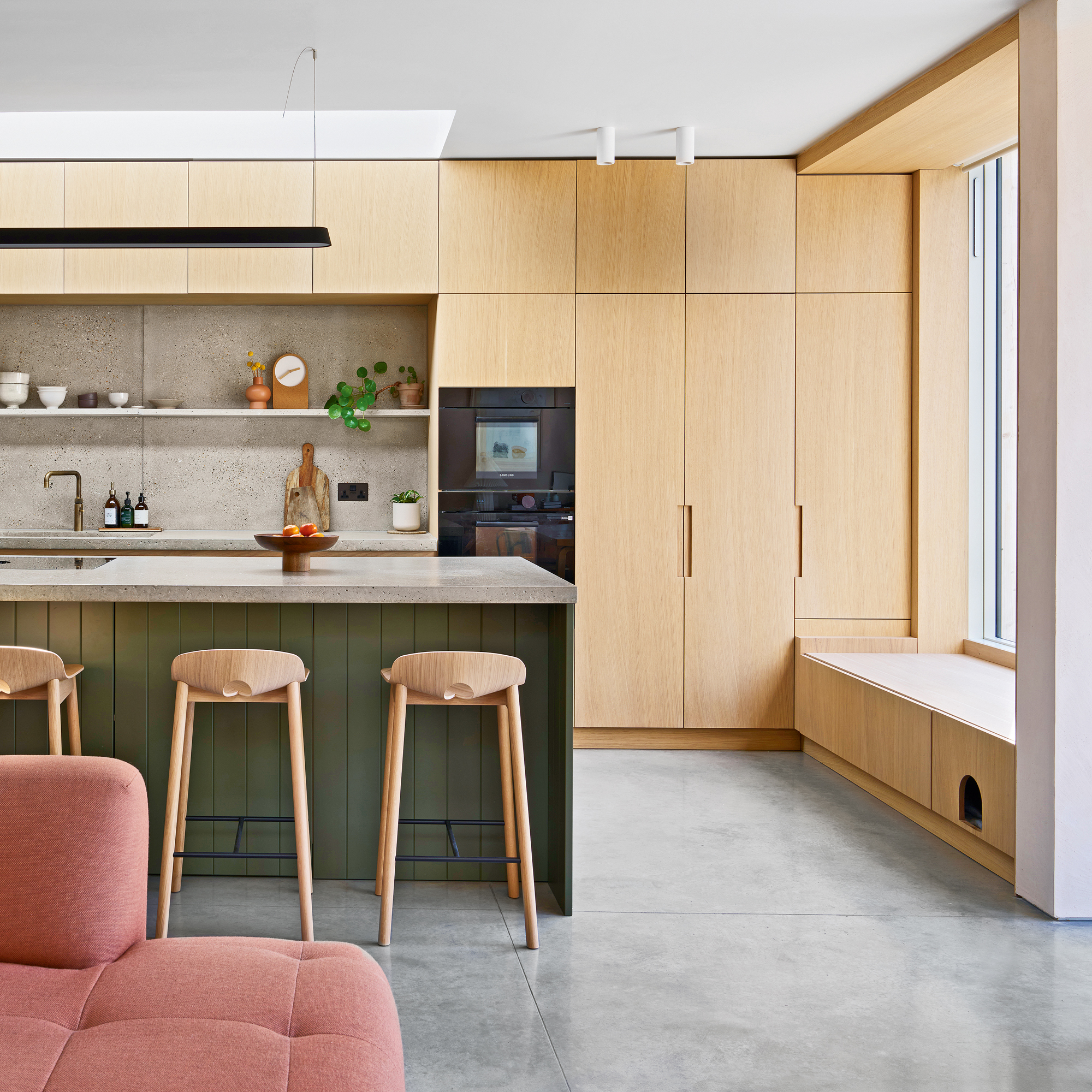 An open kitchen, a living area with a dark pink sofa and a large kitchen island with bar hops and a window seat with catblap door