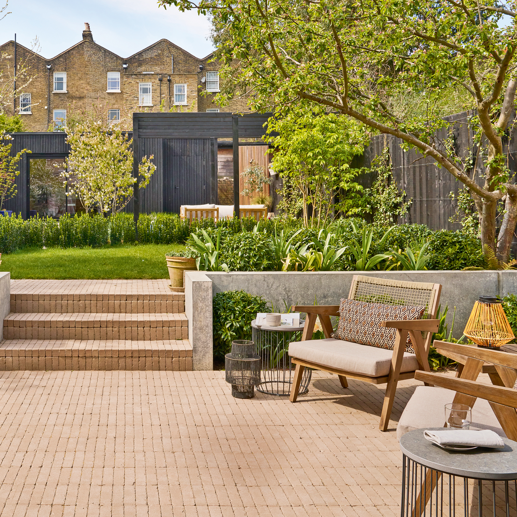 A large coil garden with a black time at the end, trees and lawn in the middle and a brick terrace in the foreground with a seating area