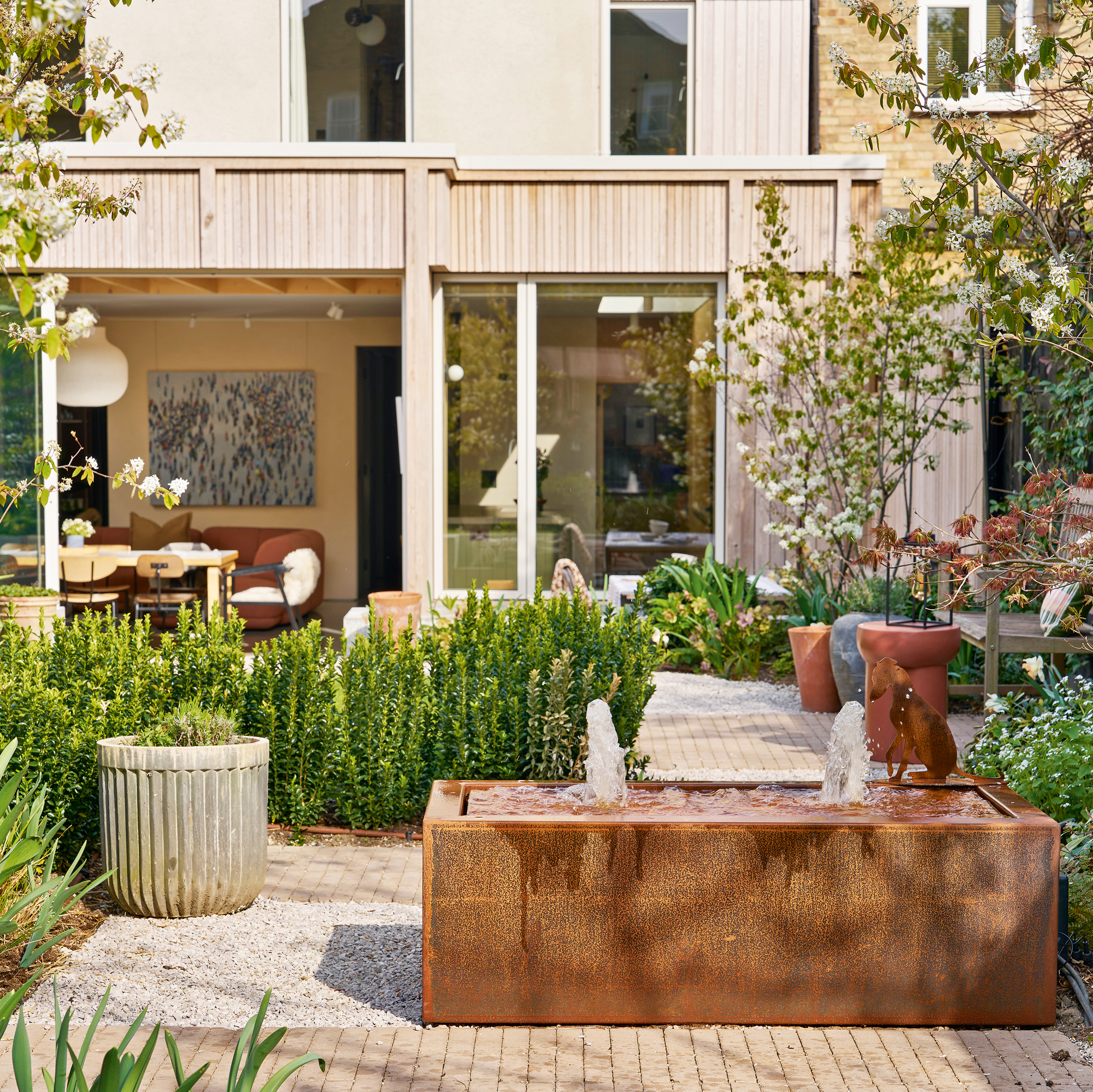 The back of a property with a large garden with a corten steel water, brick paving and many trees and plants